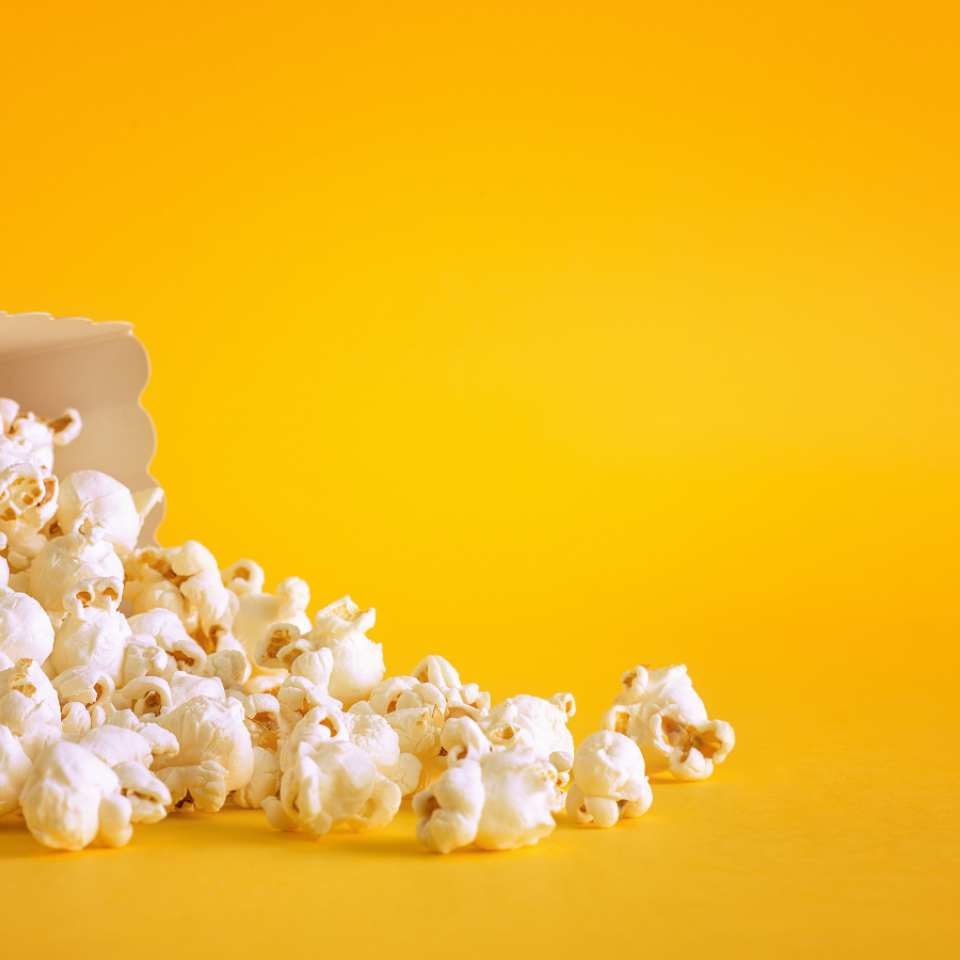 Bild: Eine rot-weiß-gestreifte Popcorntüte liegt umgekippt vor dunkelgelbem Hintergrund. Das Popcorn ist teilw. herausgefallen.
