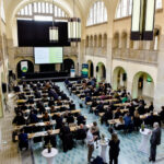 HFDcon 2024: Teilnehmende einer Konferenz sitzen an 18 Tischreihen, die im Stadtbad Oderberger aufgestellt worden sind.