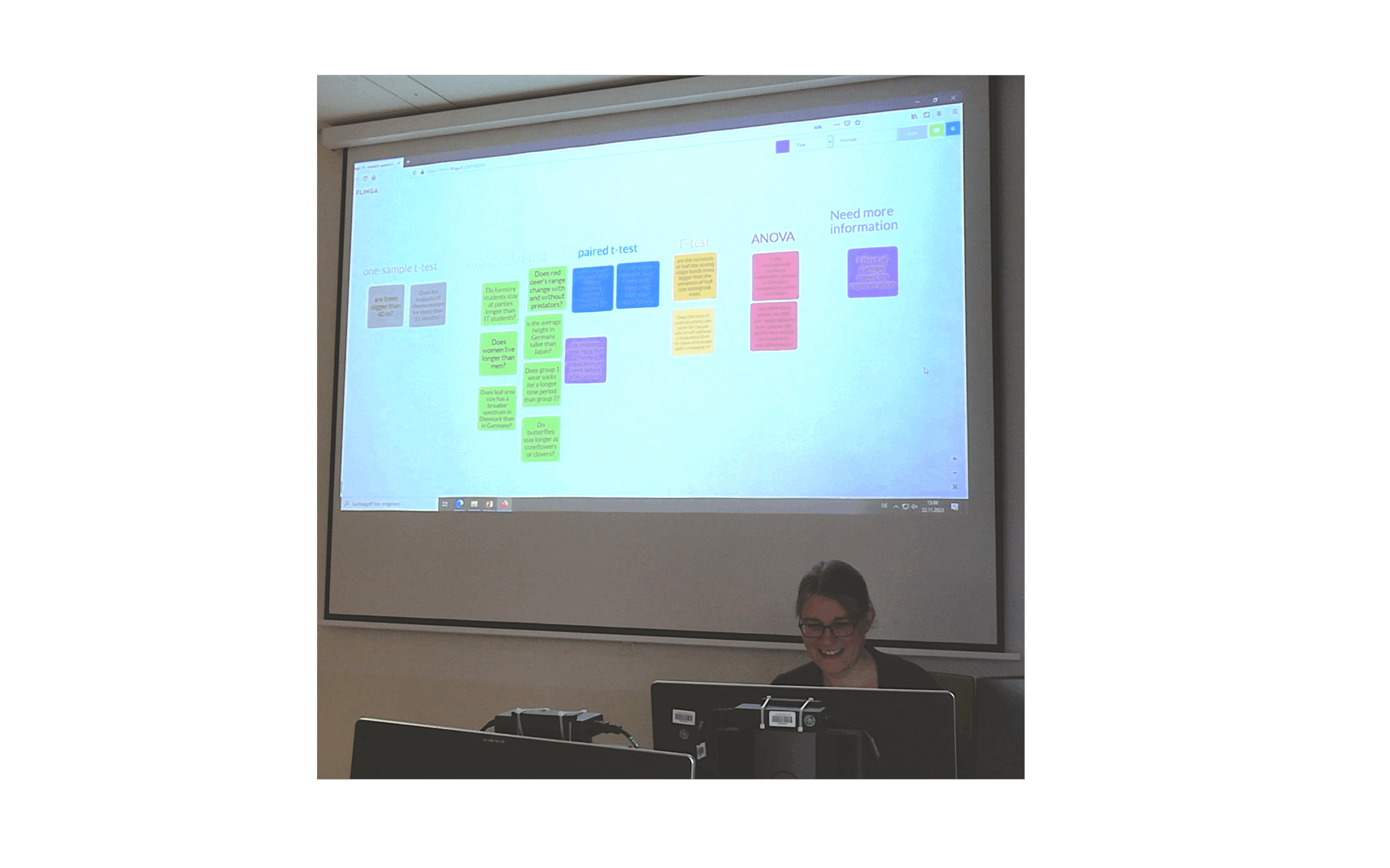 Standbild06_bearbeitet “Katrin sitzt am Rechner und bedient das Whiteboard. Sie ist fröhlich und entspannt. Hinter ihr zeigt die Projektion nach Farbe sortierte Kärtchen auf dem Board.“