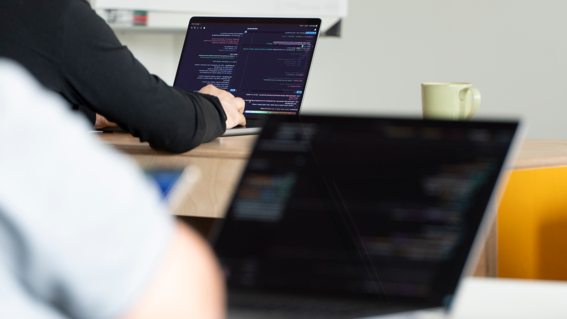 Laptop mit Code-Text, davor sieht man die Arme von Menschen, die sich auf Tische stützen.